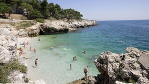 A rocky coastline with clear turquoise water, people swimming and sunbathing, and a building in the background. - Arena Verudela Beach Apartments (Pula)