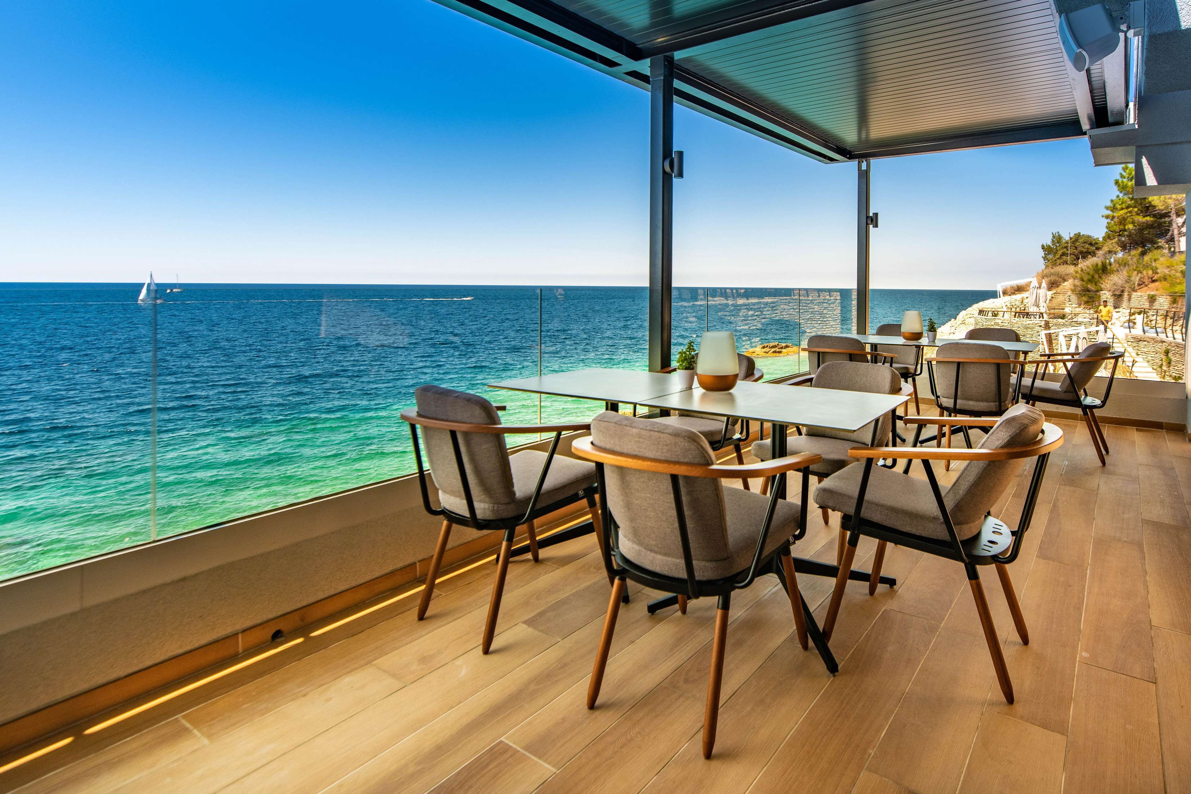 A modern outdoor dining area with a view of the sea, featuring wooden flooring, contemporary chairs, and a glass railing.