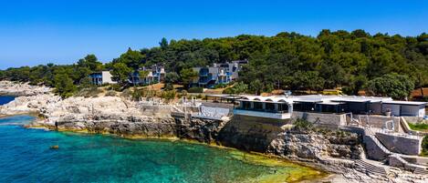 A coastal hotel with multiple buildings, a rocky shoreline, clear blue water, and a forested hill in the background.