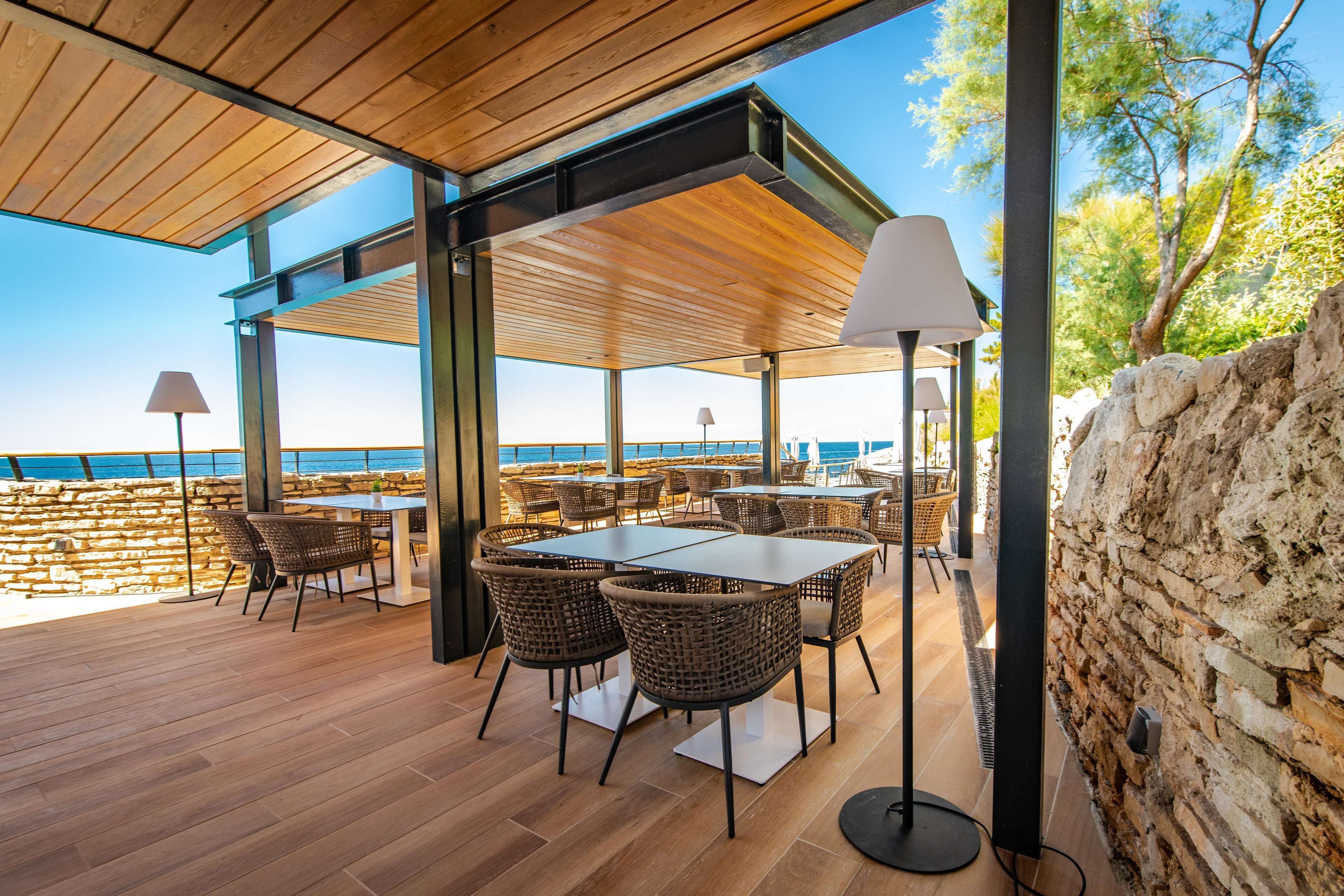 Un espace extérieur aménagé pour les repas, avec des tables et des chaises, un mur en pierre et une vue sur la mer.