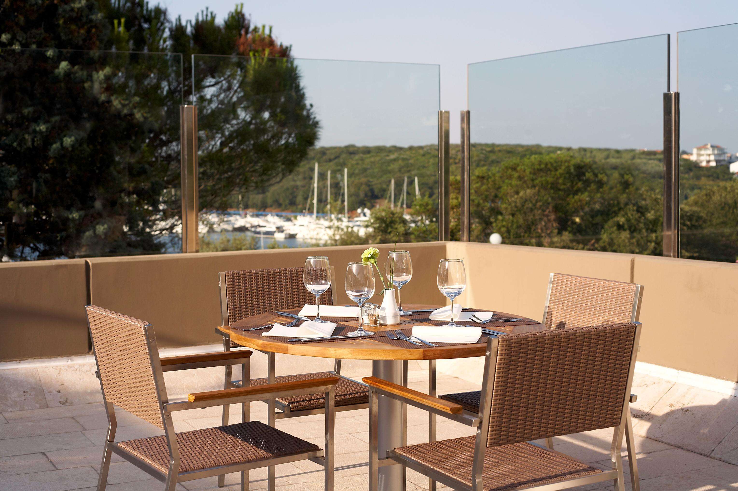 A round table with four wicker chairs, set for dining with wine glasses and a vase of flowers.