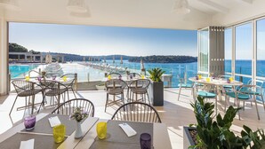 A sunlit outdoor dining area with tables and chairs, overlooking a pool and a scenic view of hills and water. - Arena Verudela Beach Apartments (Pula)