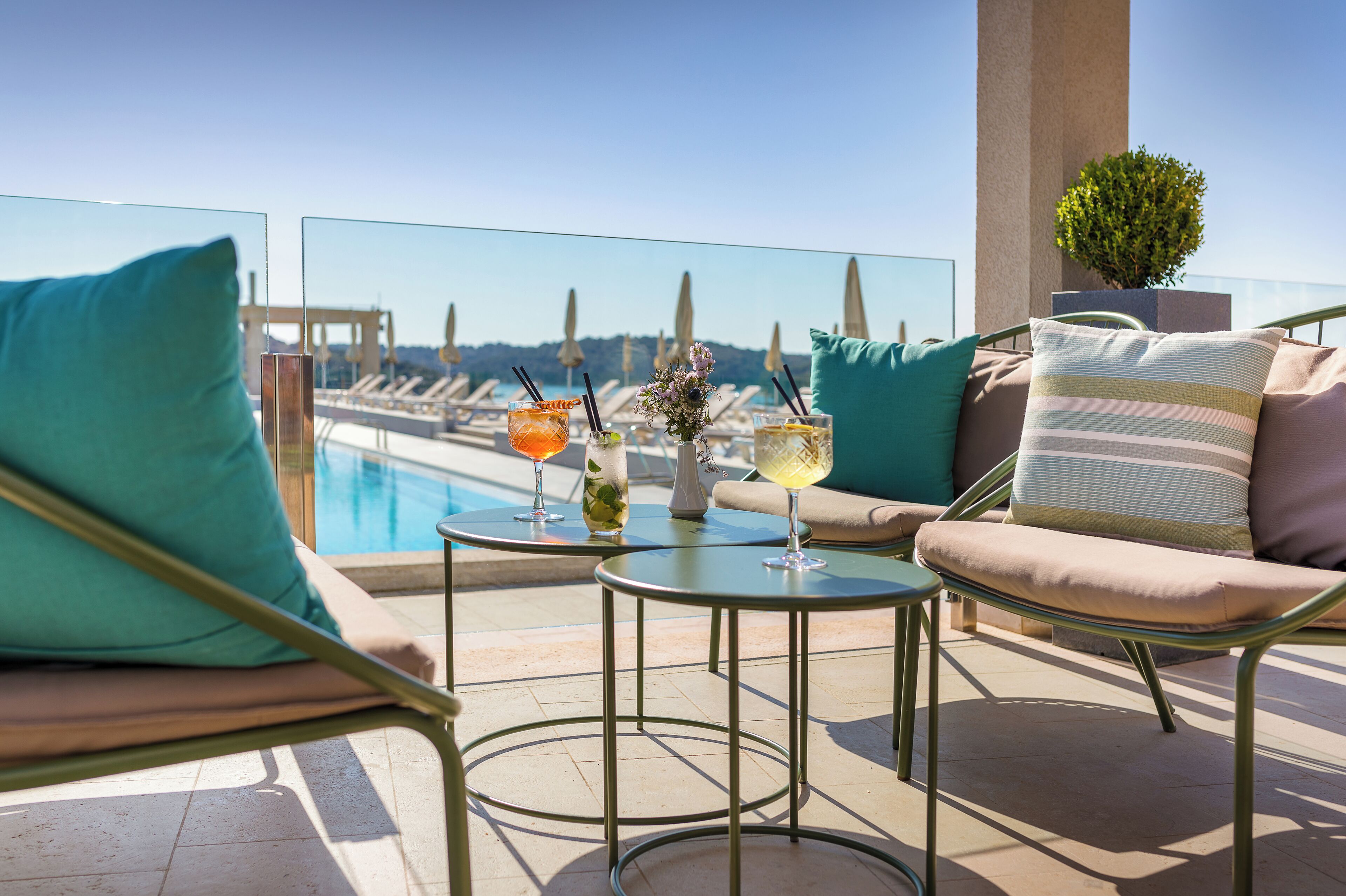 Eine Dachterrasse mit Blick auf einen Pool, zwei runde Tische mit Getränken und Lounge-Stühle mit türkisfarbenen Kissen.