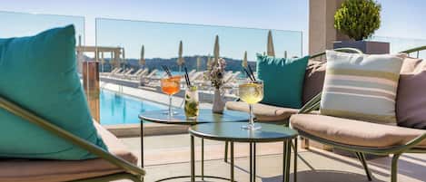 A rooftop terrace with a pool view, two round tables with drinks, and lounge chairs with teal cushions.