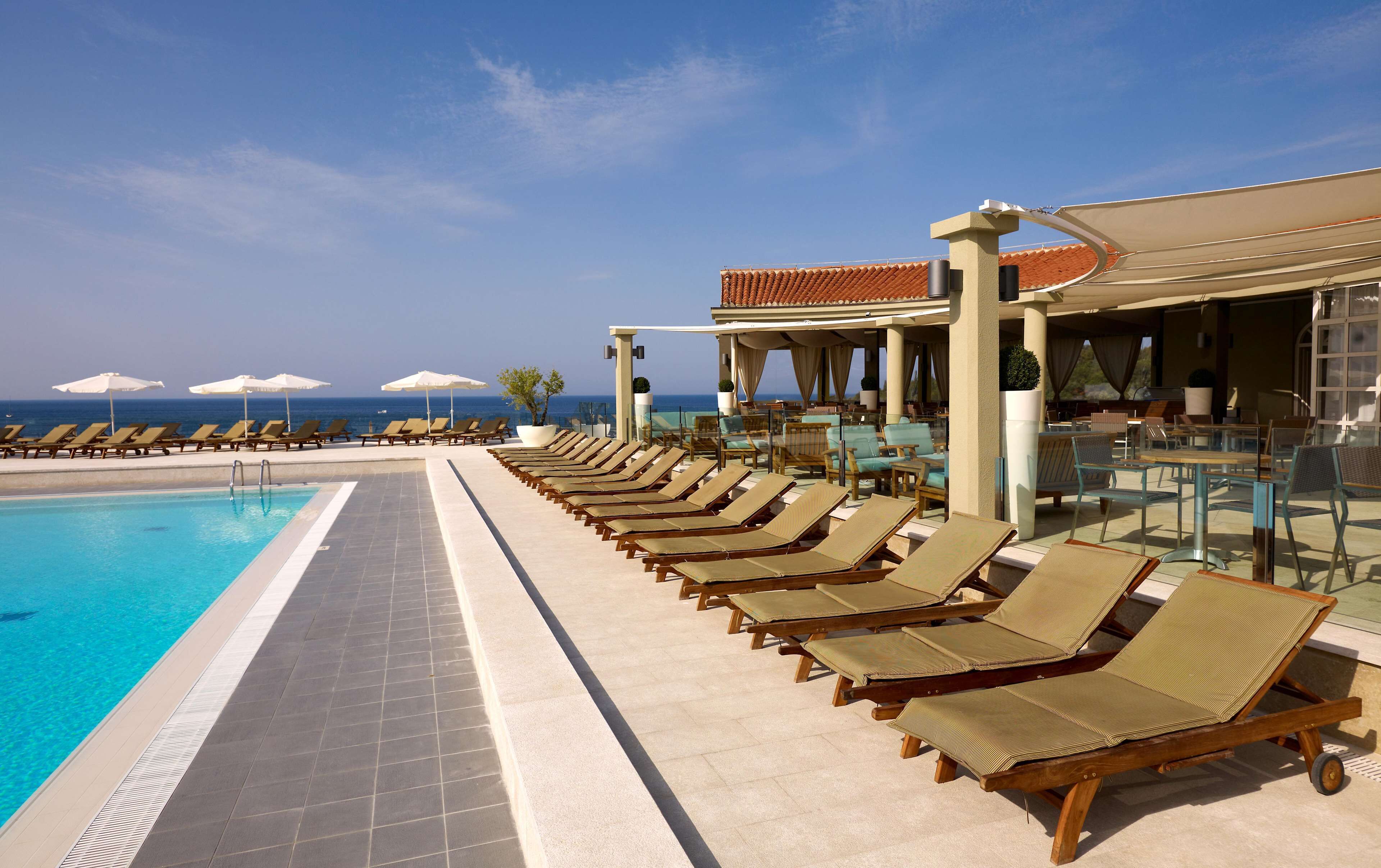 A poolside area with sun loungers and umbrellas, a bar, and a view of the ocean.
