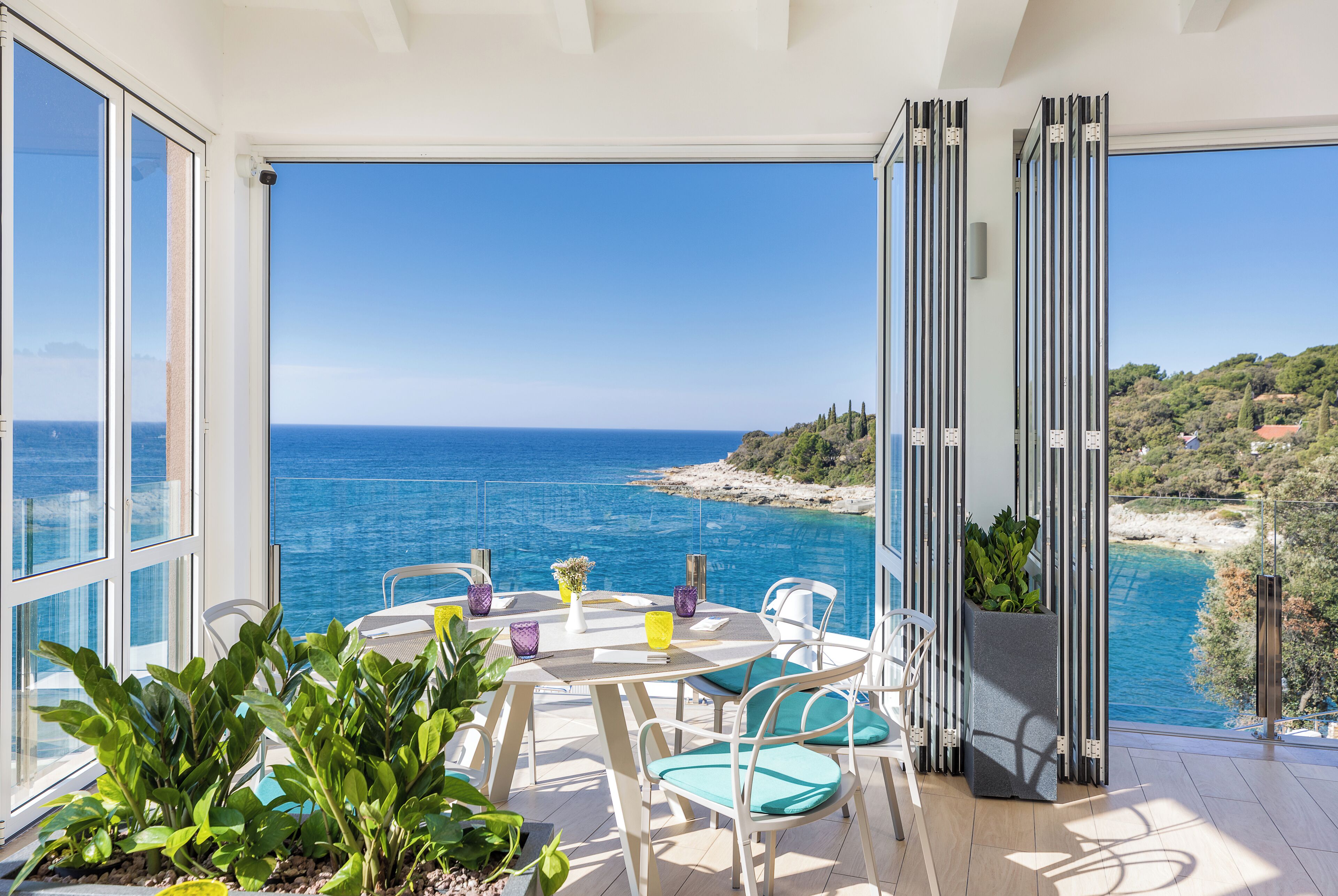 A balcony with a view of the sea, a table with chairs, and potted plants.