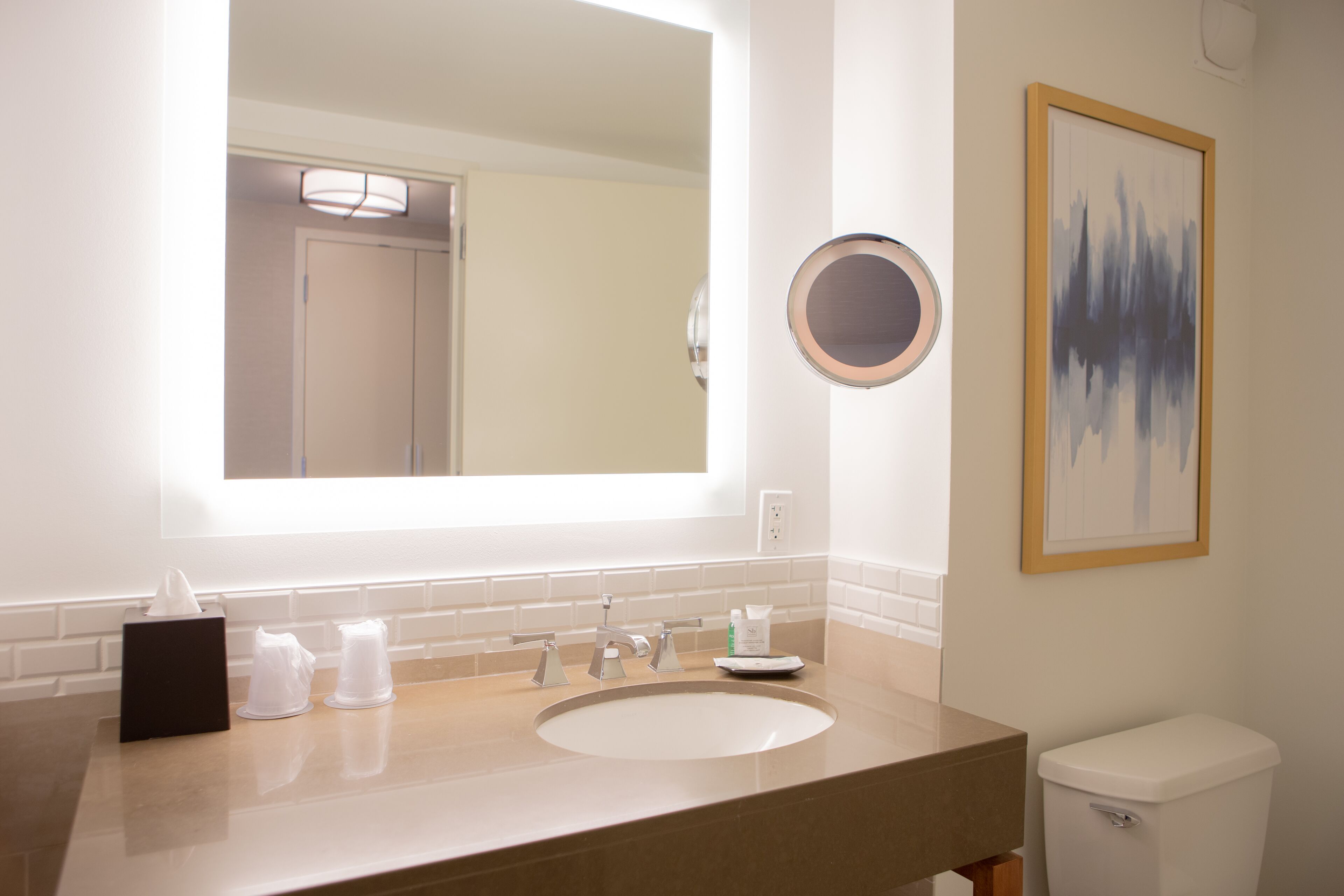 A bathroom with a large mirror, a sink, a toilet, and a framed abstract painting on the wall.