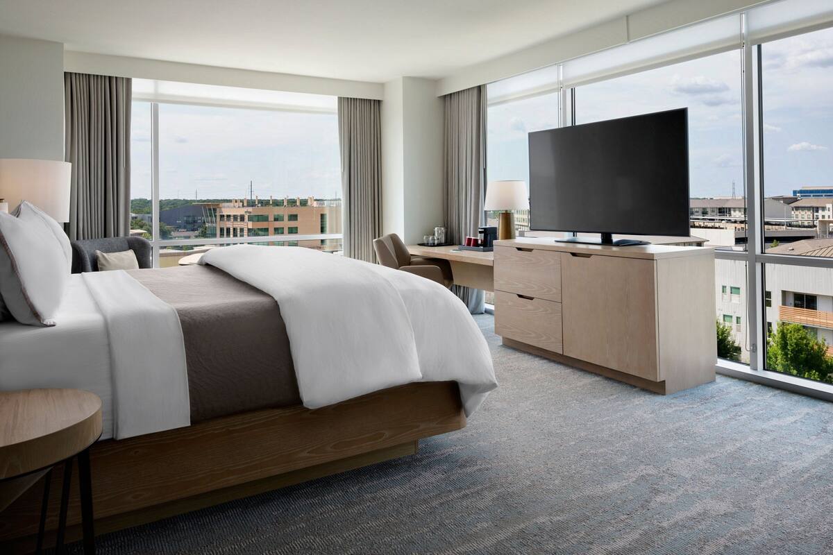 A modern hotel room with a large bed, a desk with a flat-screen TV, and a view of the cityscape through large windows.
