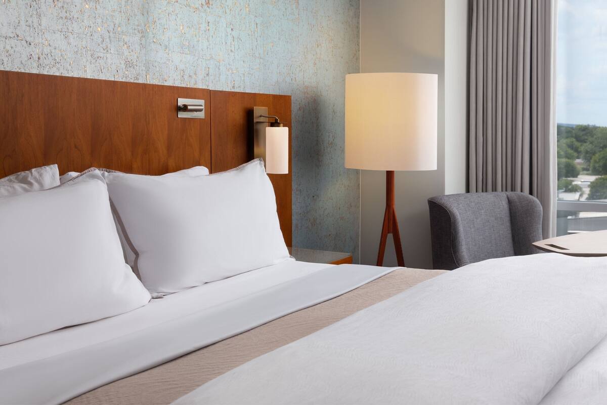 A hotel room with a large bed, a wooden headboard, a nightstand with a lamp, a grey armchair, and a view of greenery through the window.