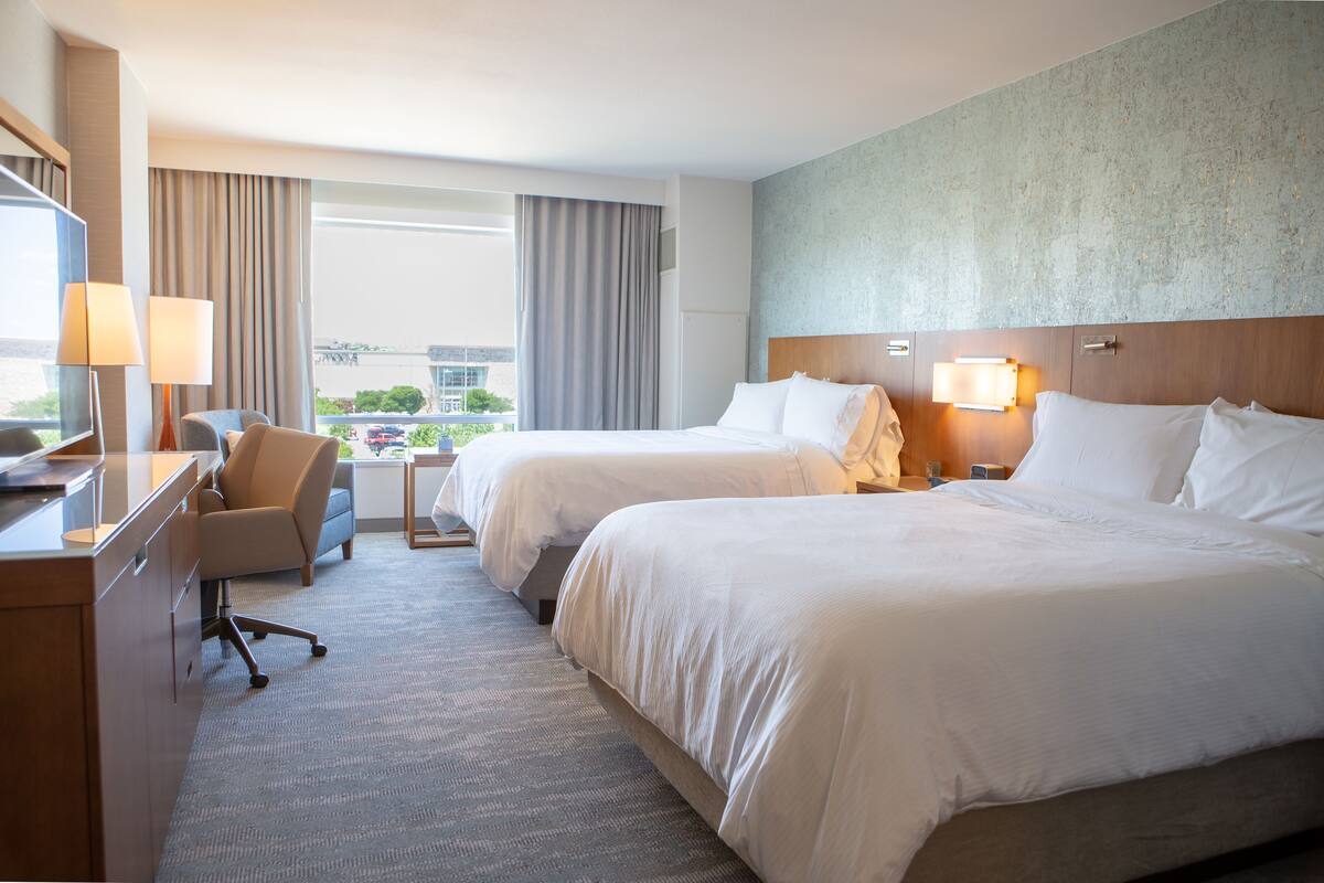 A hotel room with two beds, a desk, a chair, and a view of the city.