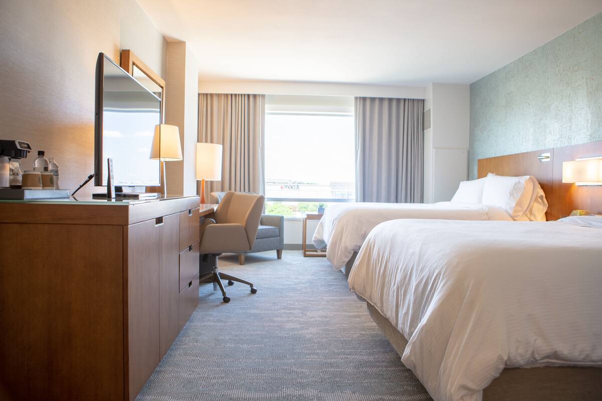 A hotel room with a desk, two beds, a chair, a large mirror, and a view of the outdoors.