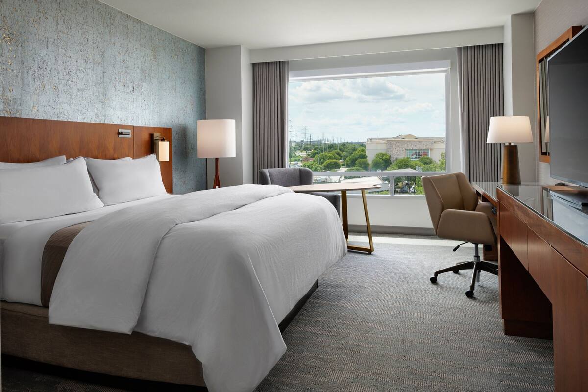 A hotel room with a large bed, a desk, a chair, a TV, and a view of the city through the window.