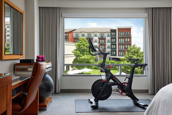 A hotel room with a view of a building, a desk with a chair, a black exercise bike, and a yoga mat.