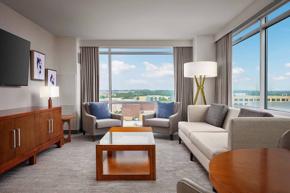 A modern hotel room with a large window, a flat-screen TV, a sofa, a coffee table, and a side table.