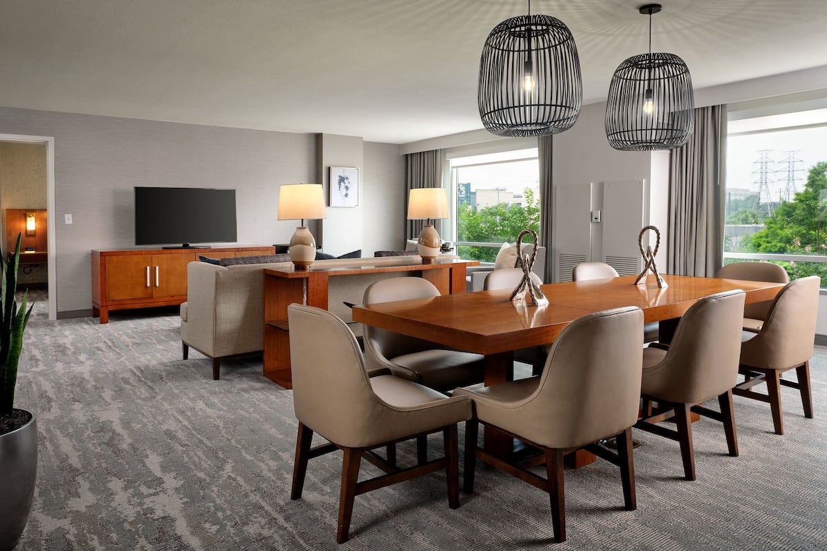 A modern hotel room with a wooden dining table, six chairs, a TV, a minibar, and a large window with a city view.