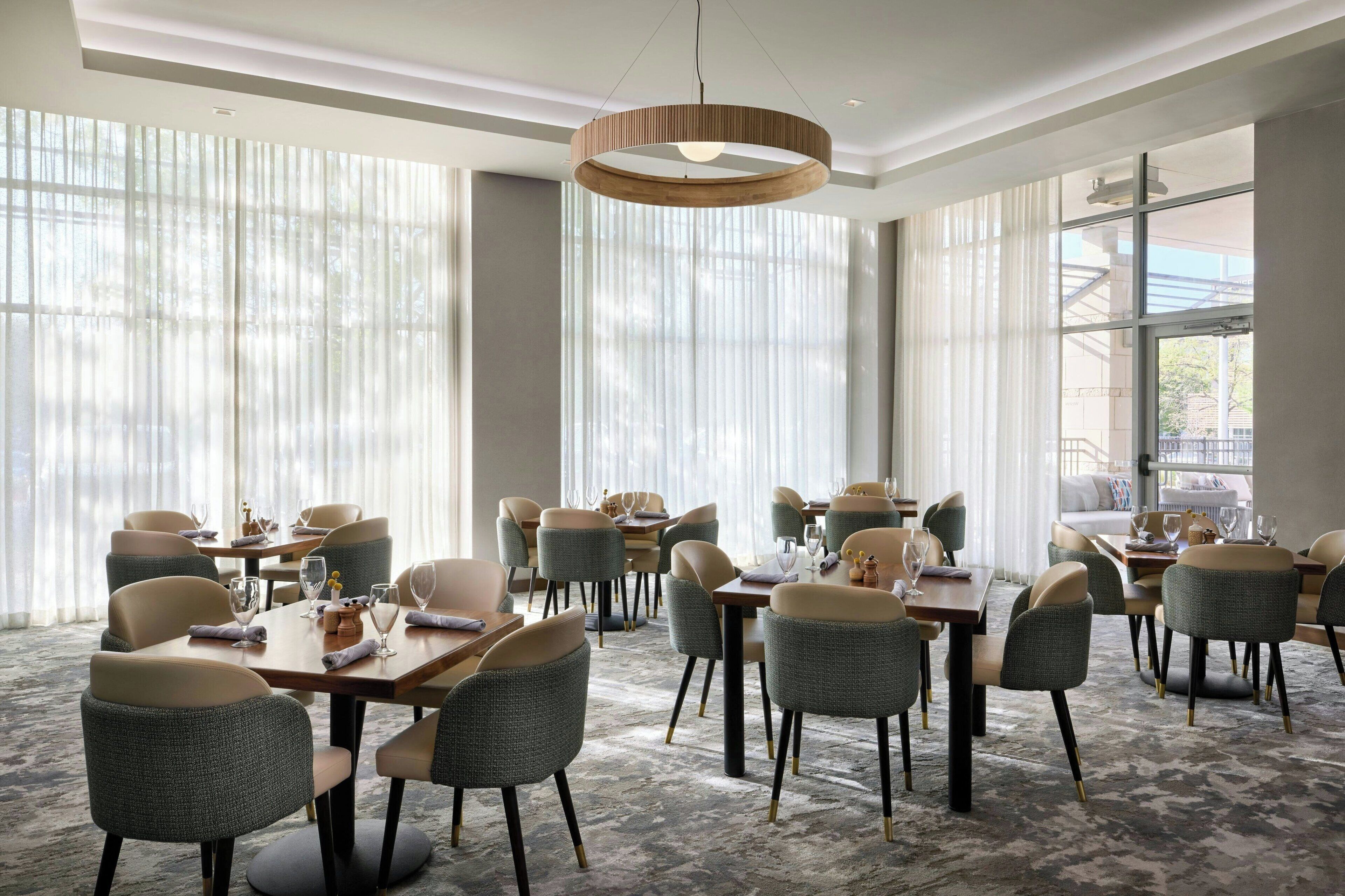 Un'area pranzo moderna con tavoli rotondi e sedie imbottite, ampie finestre con tende leggere e un lampadario centrale.