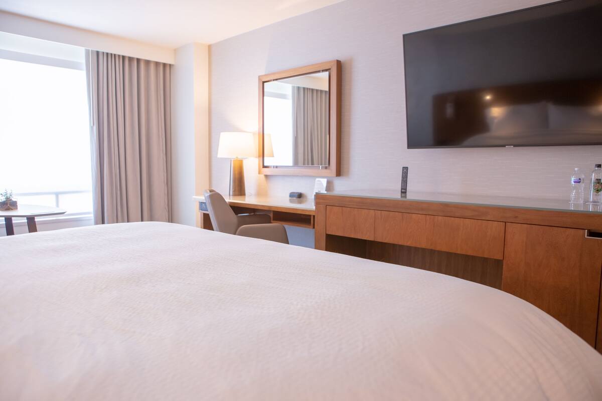A hotel room with a large bed, a desk with a chair, a flat-screen TV mounted on the wall, a mirror, and a window with curtains.