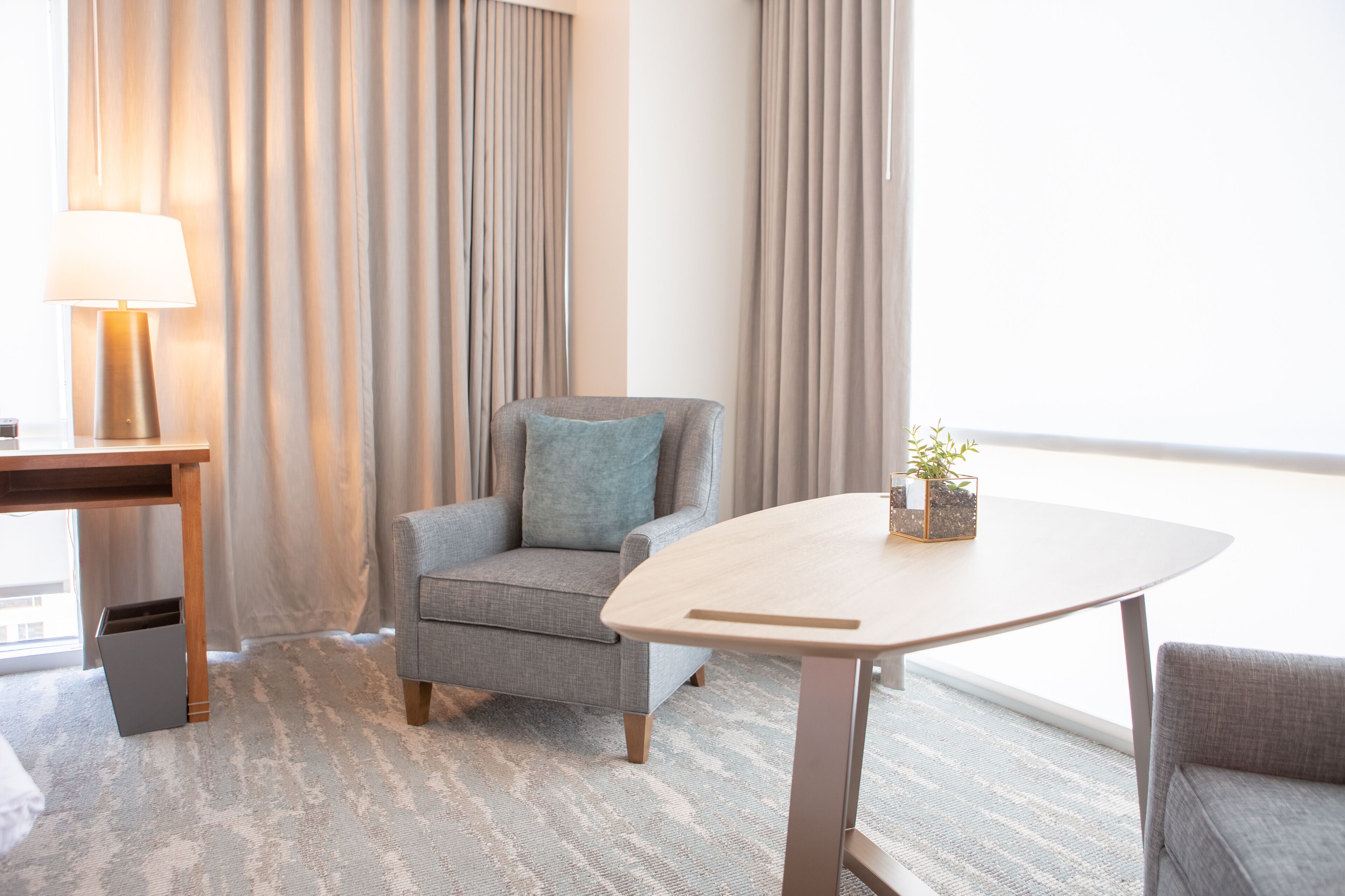 Une chambre d’hôtel moderne avec un fauteuil gris, une table en bois avec une lampe et une vue sur la ville grâce à de grandes fenêtres.