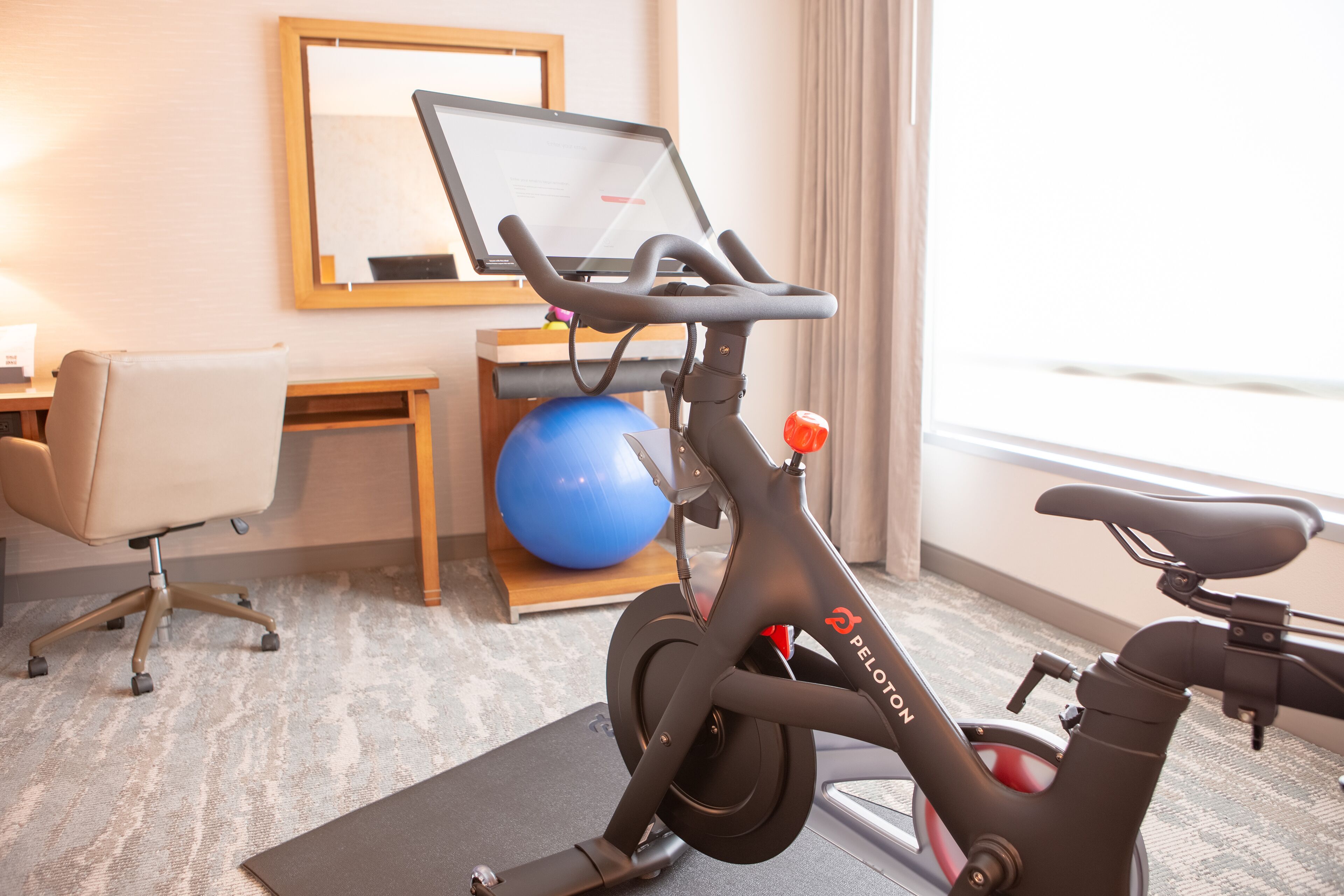A Peloton stationary bike in a hotel room with a desk, chair, exercise ball, and a window with curtains.