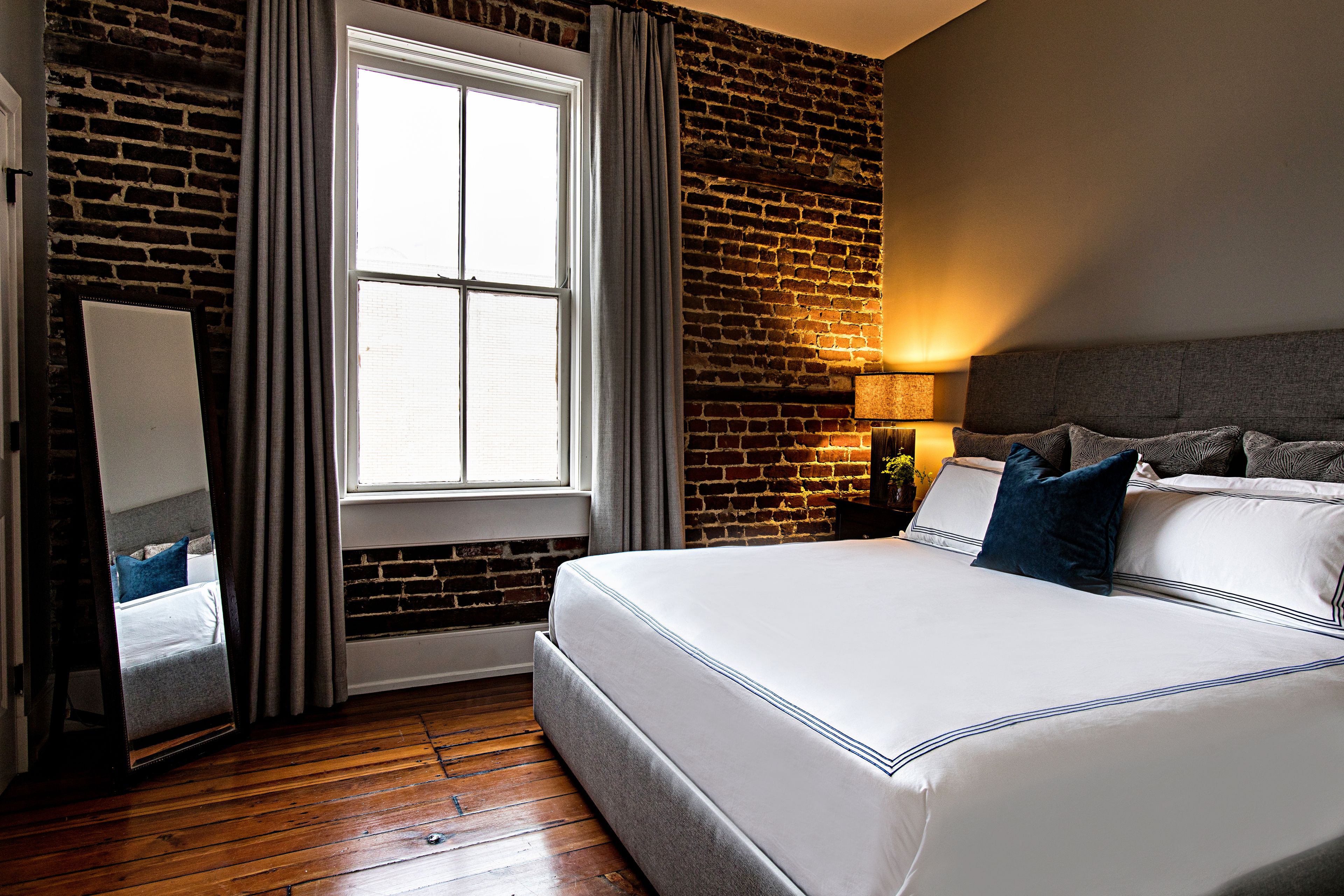 A hotel room with a brick wall, a large window with curtains, a bed with white bedding, and a nightstand with a lamp.