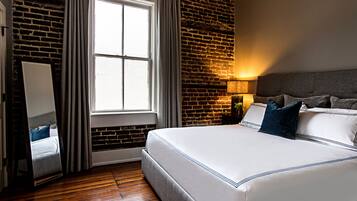 A hotel room with a brick wall, a large window with curtains, a bed with white bedding, and a nightstand with a lamp.