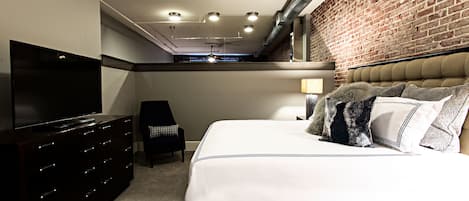 Un dormitorio con una cama, televisor, cómoda, silla y pared de ladrillo.