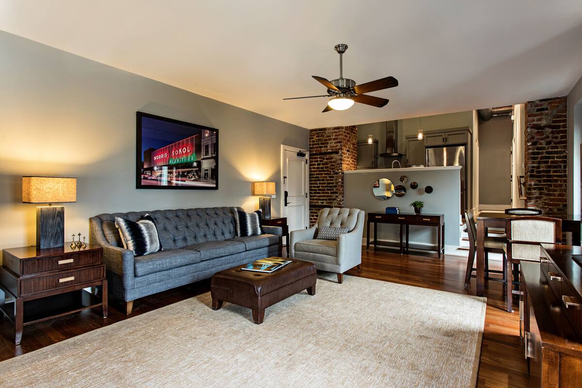 A living room with a sofa, armchair, ottoman, and coffee table. There is a ceiling fan, a lamp, and a framed picture on the wall. A kitchen is visible in the background.
