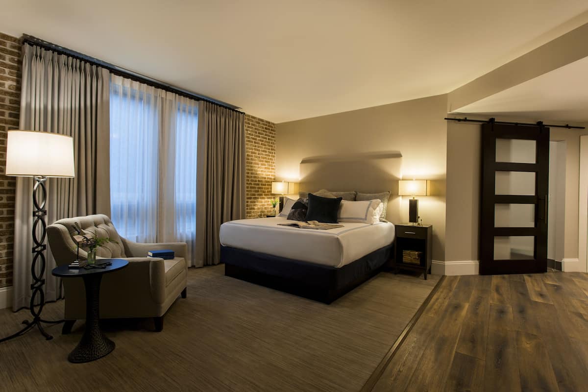 A hotel room with a bed, a chair, a table, and a lamp.
