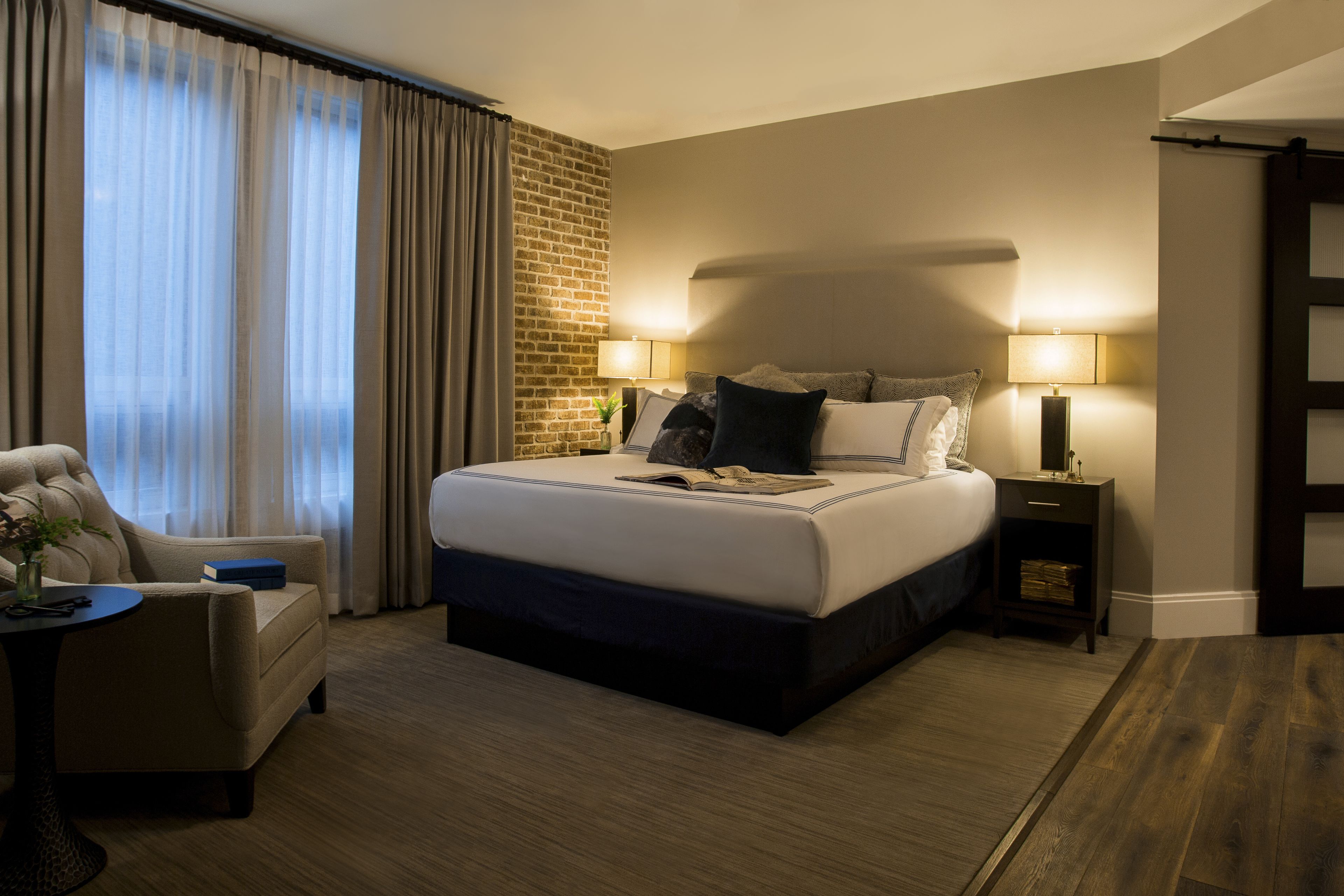A hotel room with a large bed, two bedside tables with lamps, a chair, and a view of the city through large windows.