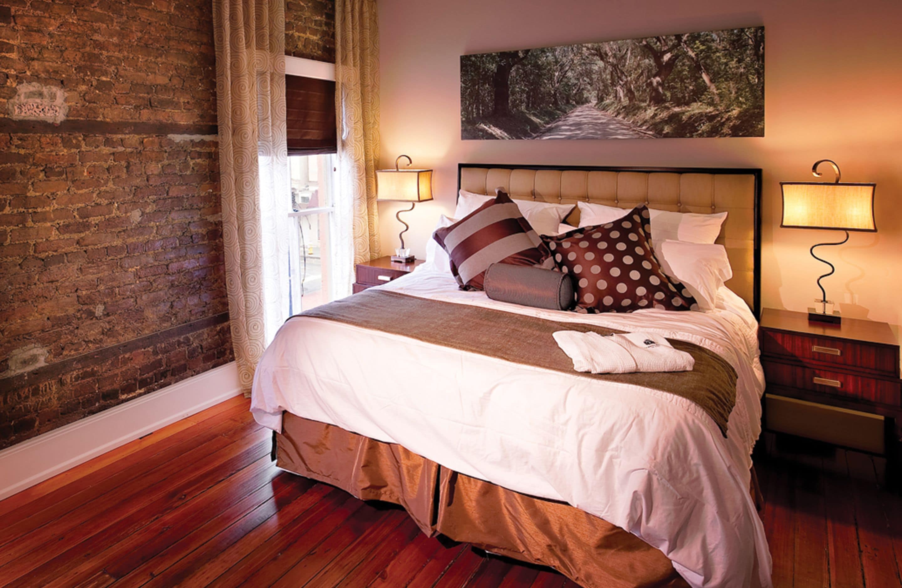 A hotel room with a brick wall, a large bed with multiple pillows, a wooden floor, and two bedside lamps.