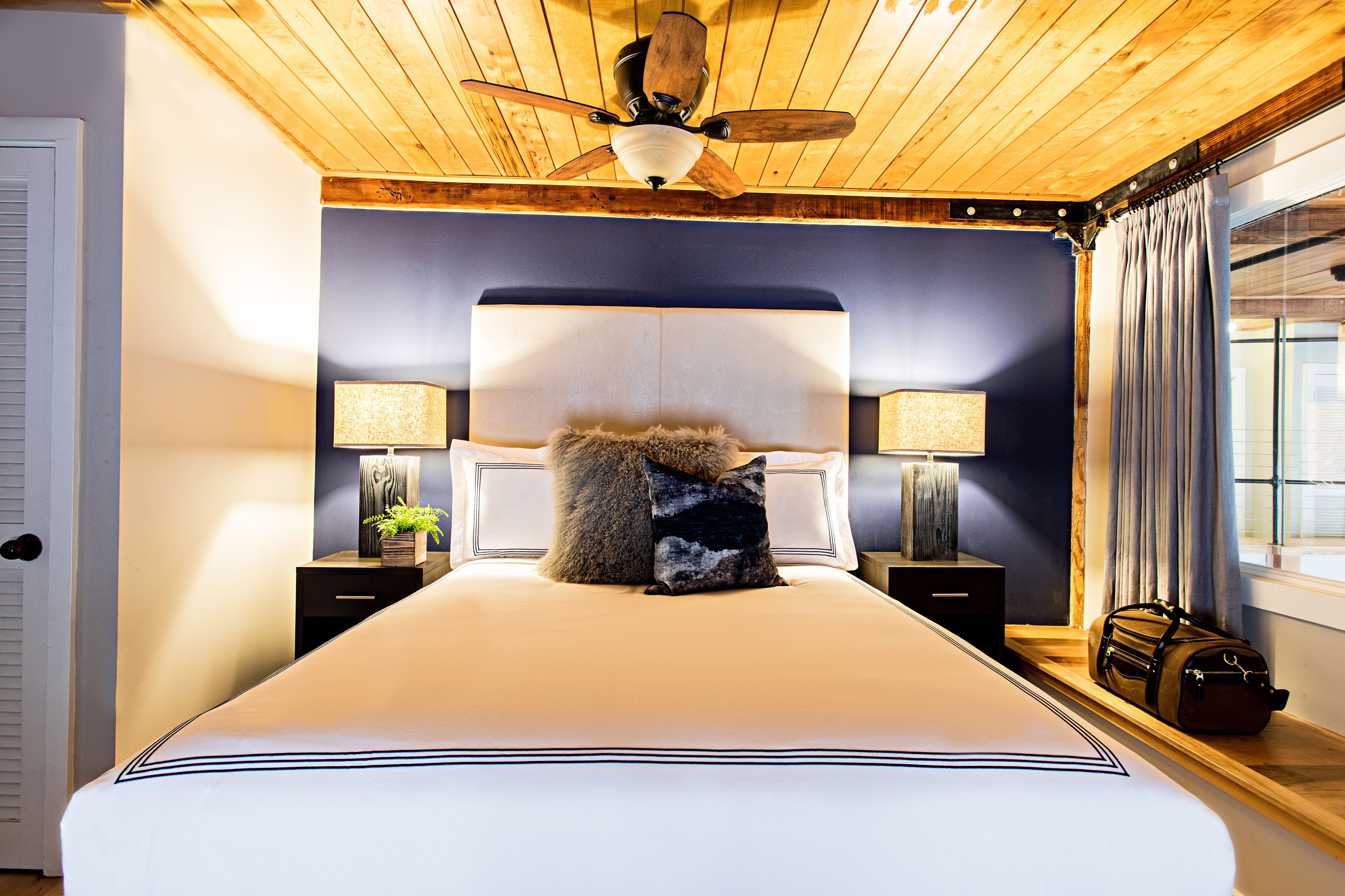 A bedroom with a large bed, wooden ceiling, two bedside lamps, a nightstand, and a window with curtains.