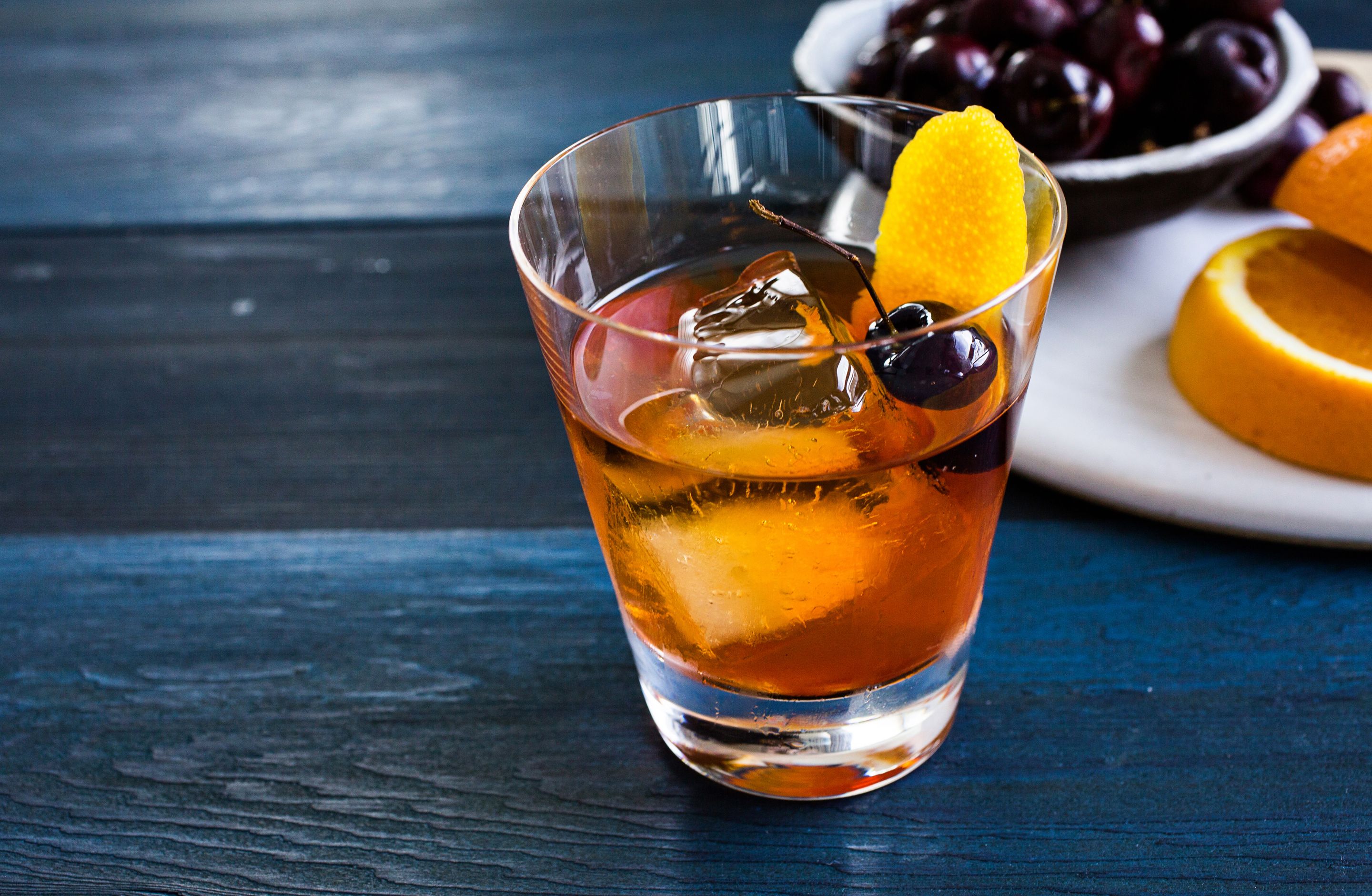 A glass of amber-colored beverage with ice cubes and a slice of orange, accompanied by a bowl of cherries and an orange half on a wooden surface.