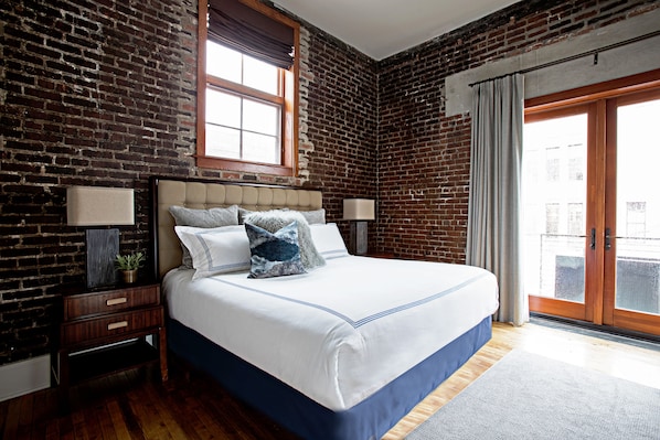 A bedroom with a brick wall, a large bed with a blue bedspread, two bedside tables with lamps, and a window with curtains.
