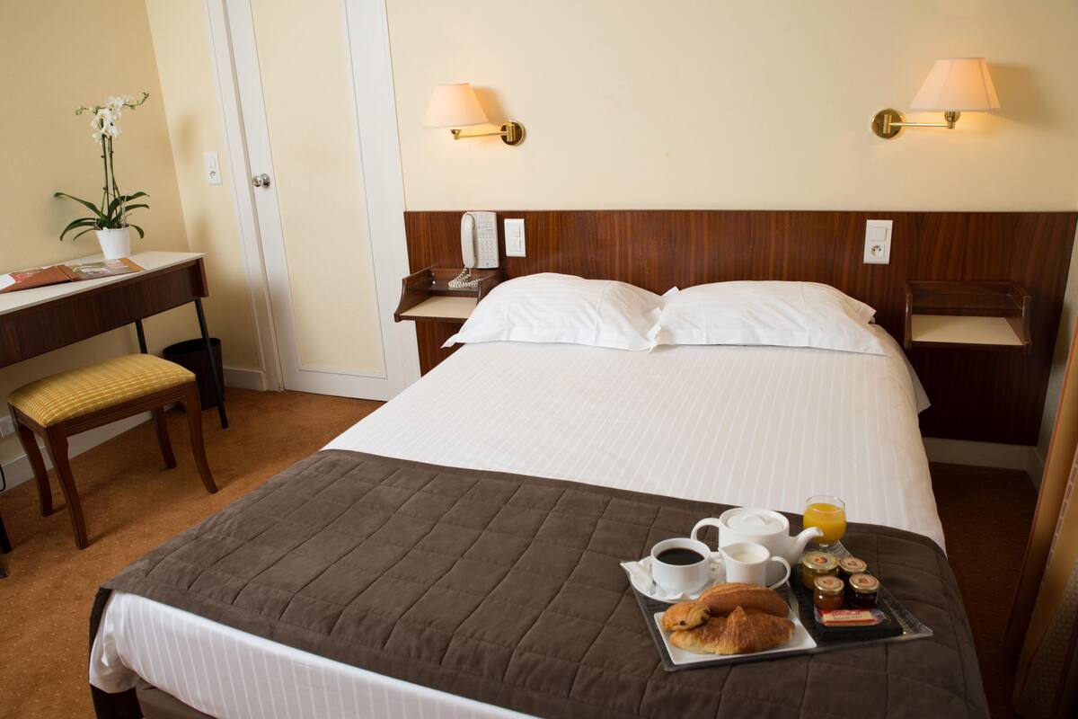 Une chambre d’hôtel avec un lit soigneusement fait, une table de chevet avec une lampe, un bureau avec une chaise et un plateau de petit-déjeuner posé sur le lit.