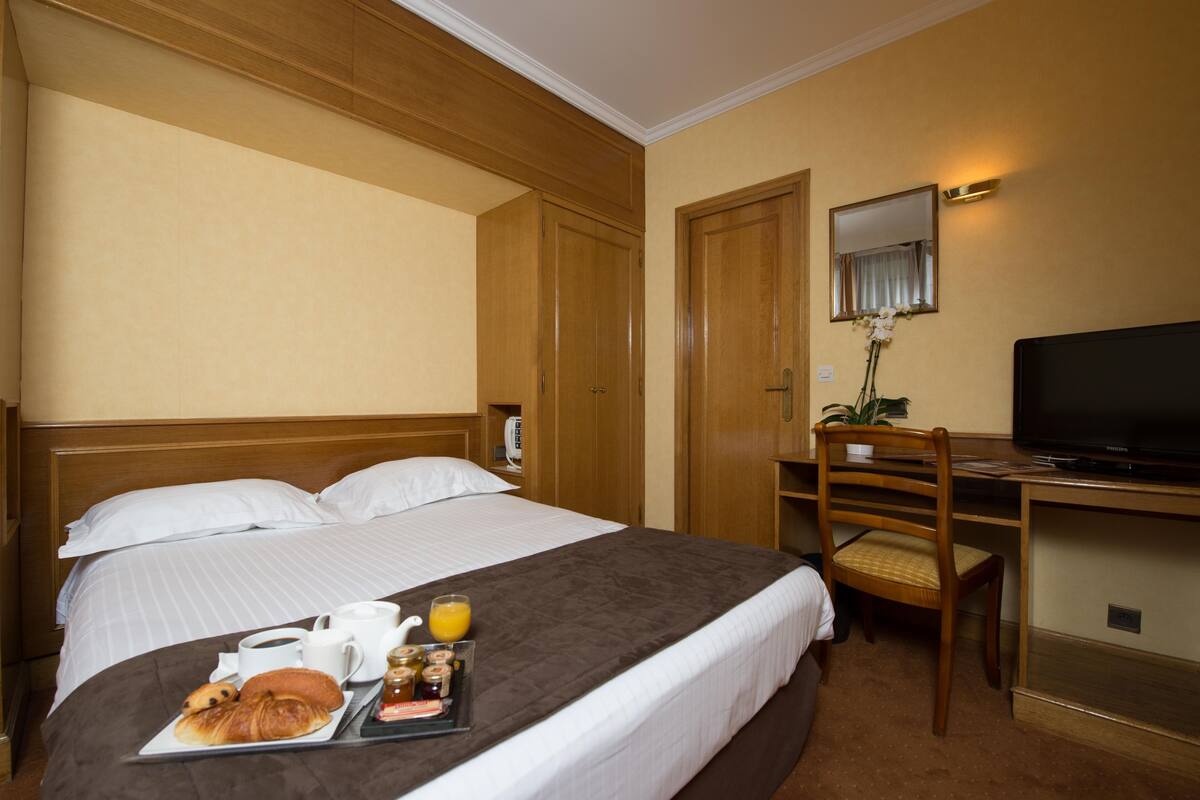 Une chambre d’hôtel comprenant un lit, un bureau avec une chaise, une télévision et un plateau de petit-déjeuner avec des croissants et du jus de fruits.