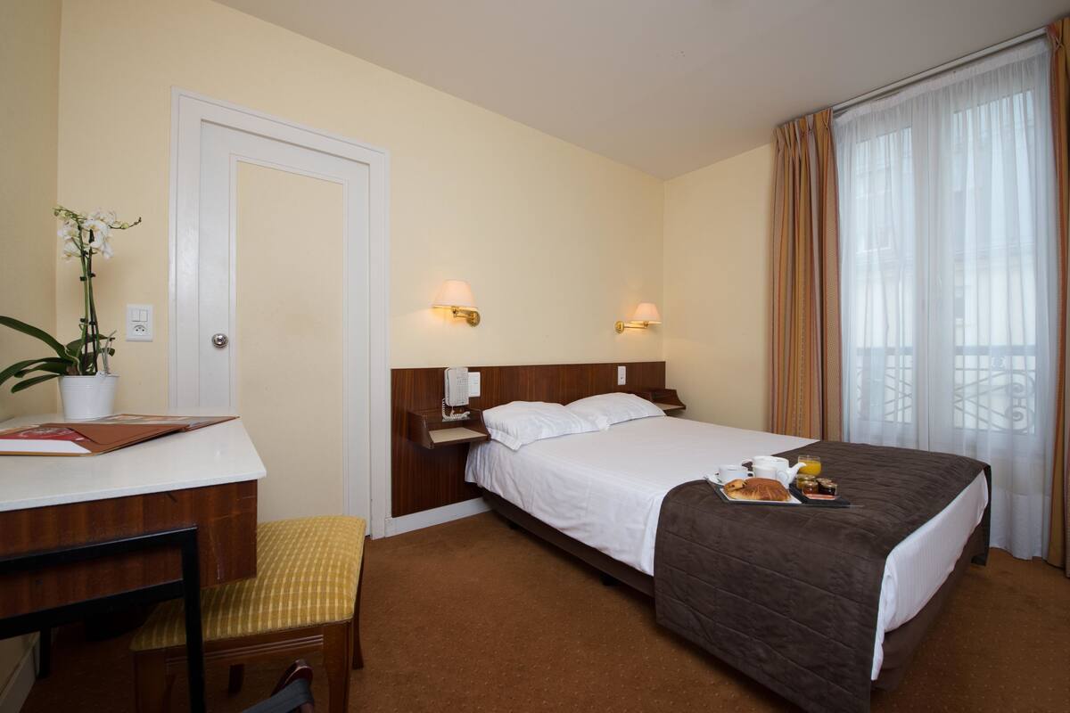 Une chambre d’hôtel comprenant un lit, des tables de chevet, un bureau, une chaise, une plante en pot et une fenêtre avec des rideaux.