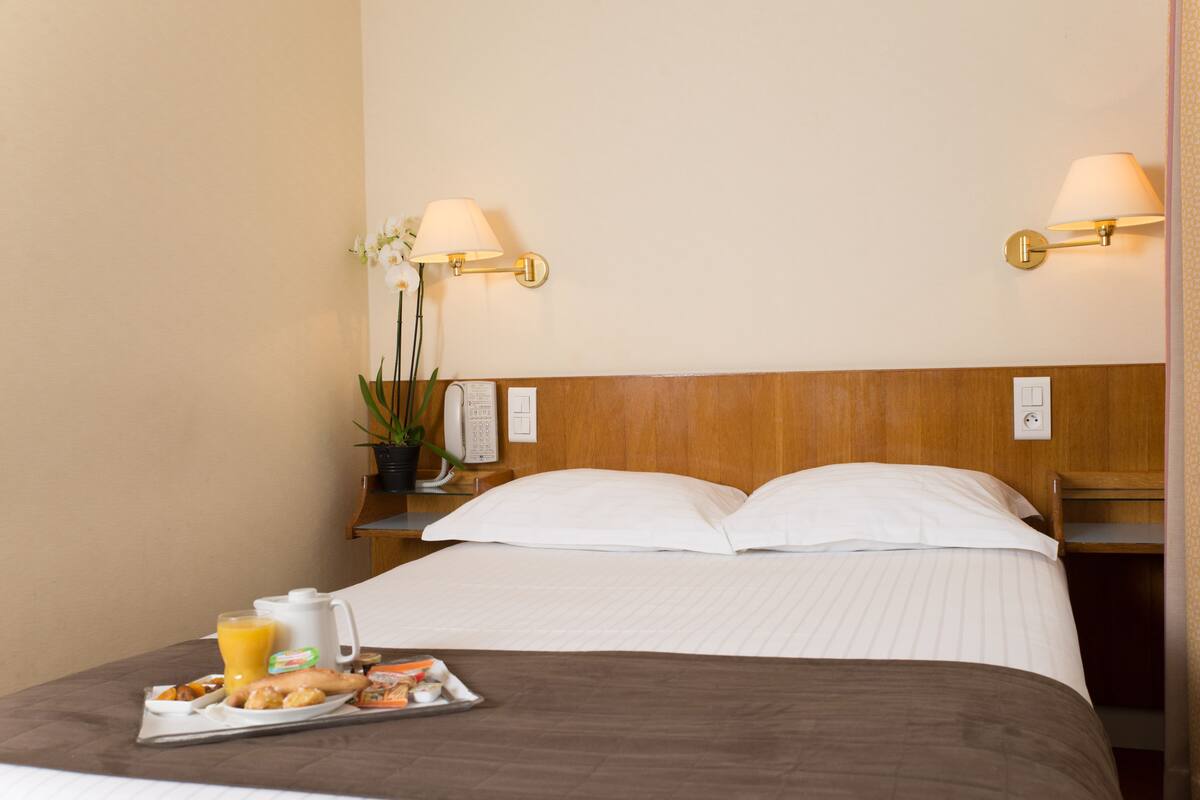 Une chambre d’hôtel avec un lit, des lampes de chevet, une table de chevet avec un téléphone, un plateau avec des articles pour le petit-déjeuner et une plante en pot.