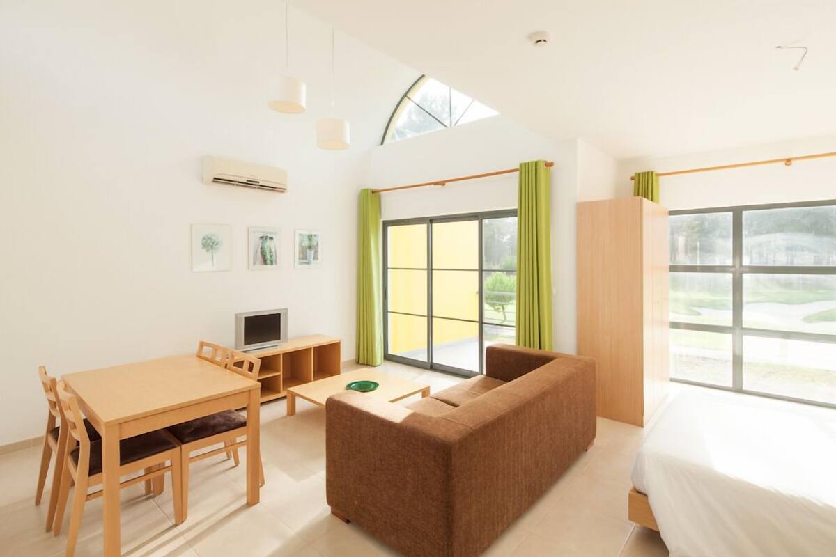A modern, well-lit room with a wooden dining table, a brown sofa, a bed, and large windows offering a view of the outdoors.