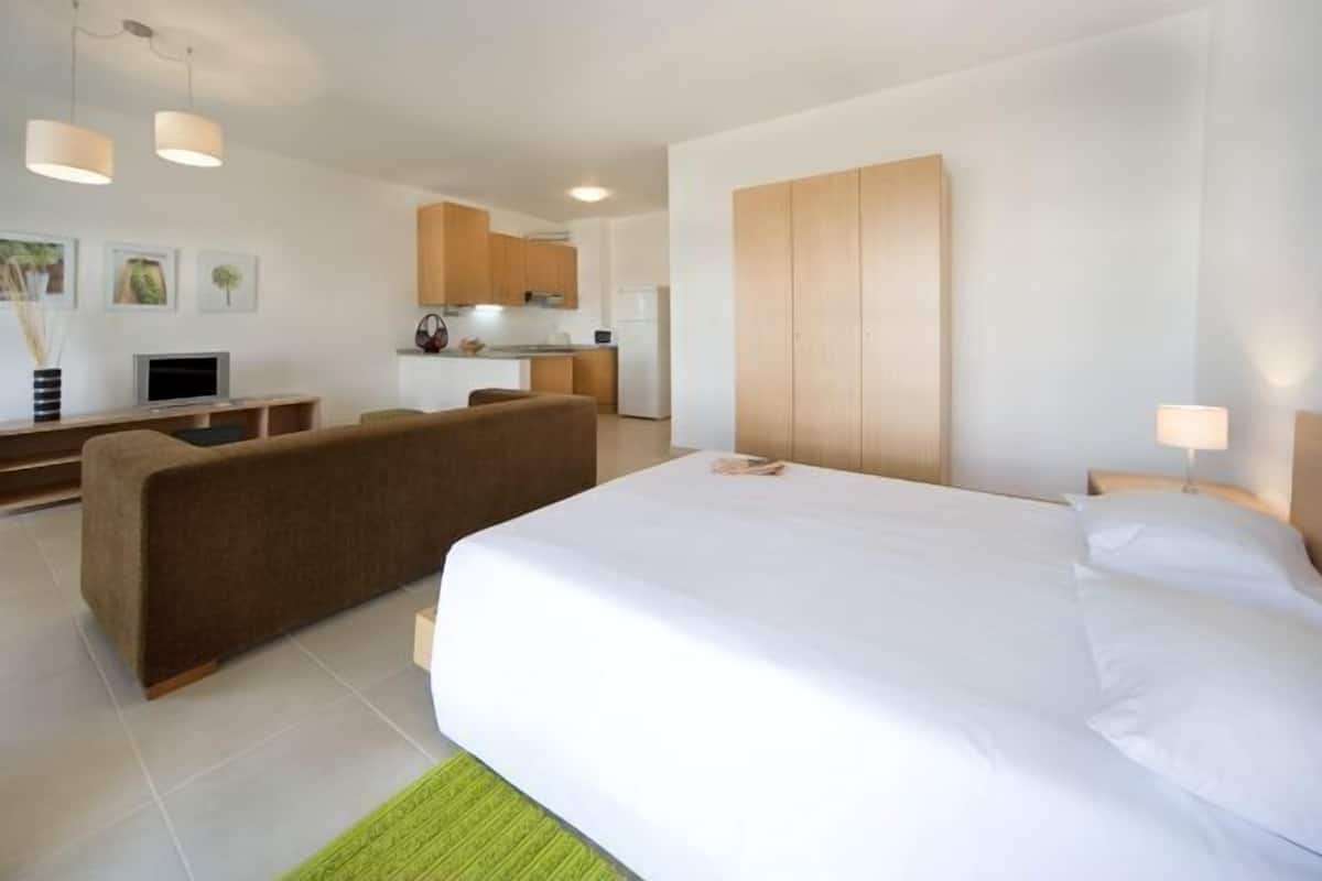 A modern hotel room with a large bed, a sofa, a small kitchen area, and a dining table.