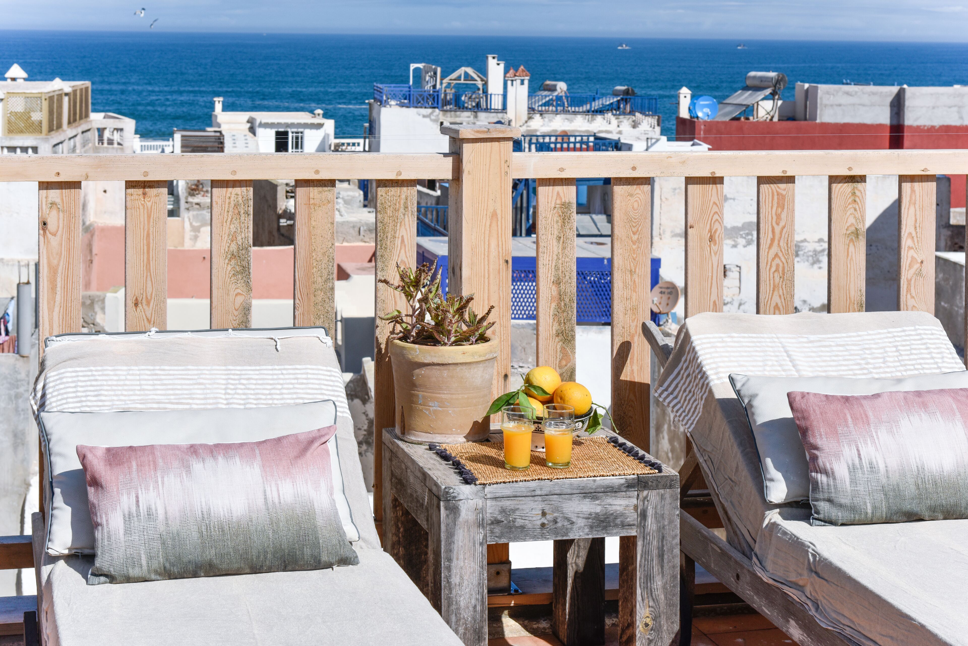 A balcony with two sunbeds, a small table with a potted plant and fruit, and a view of the sea and buildings.