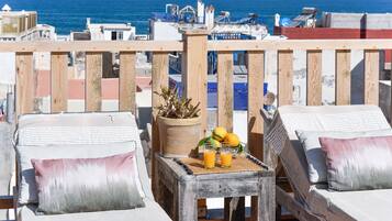 A balcony with two sunbeds, a small table with a potted plant and fruit, and a view of the sea and buildings.
