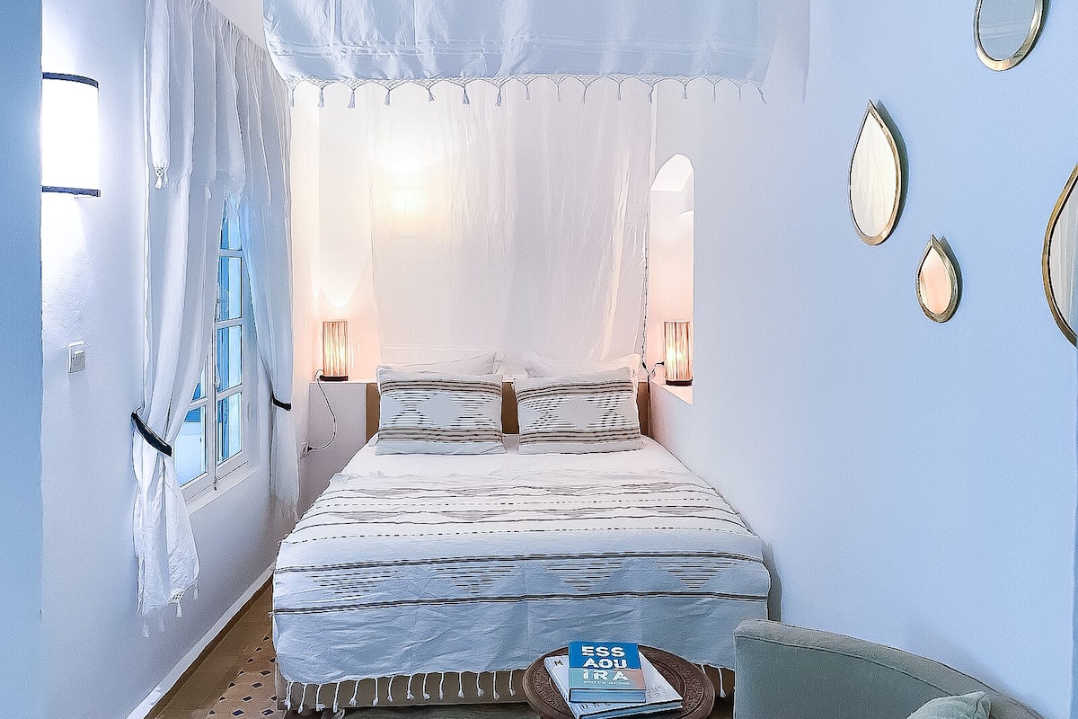 Un dormitorio con una cama, dos lámparas de noche, un ventanal con cortinas y una pequeña mesa con libros.