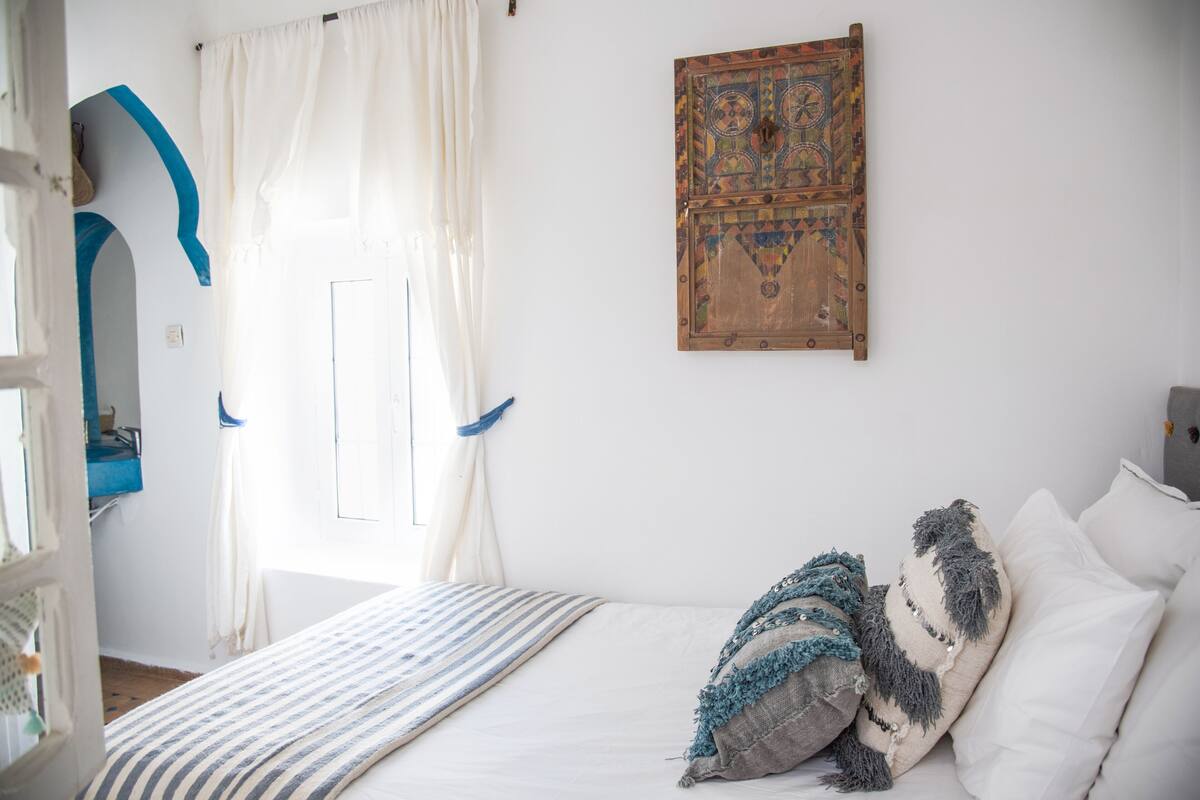 Um quarto com cama, janela com cortinas, painel decorativo fixado na parede e um arco pintado de azul.