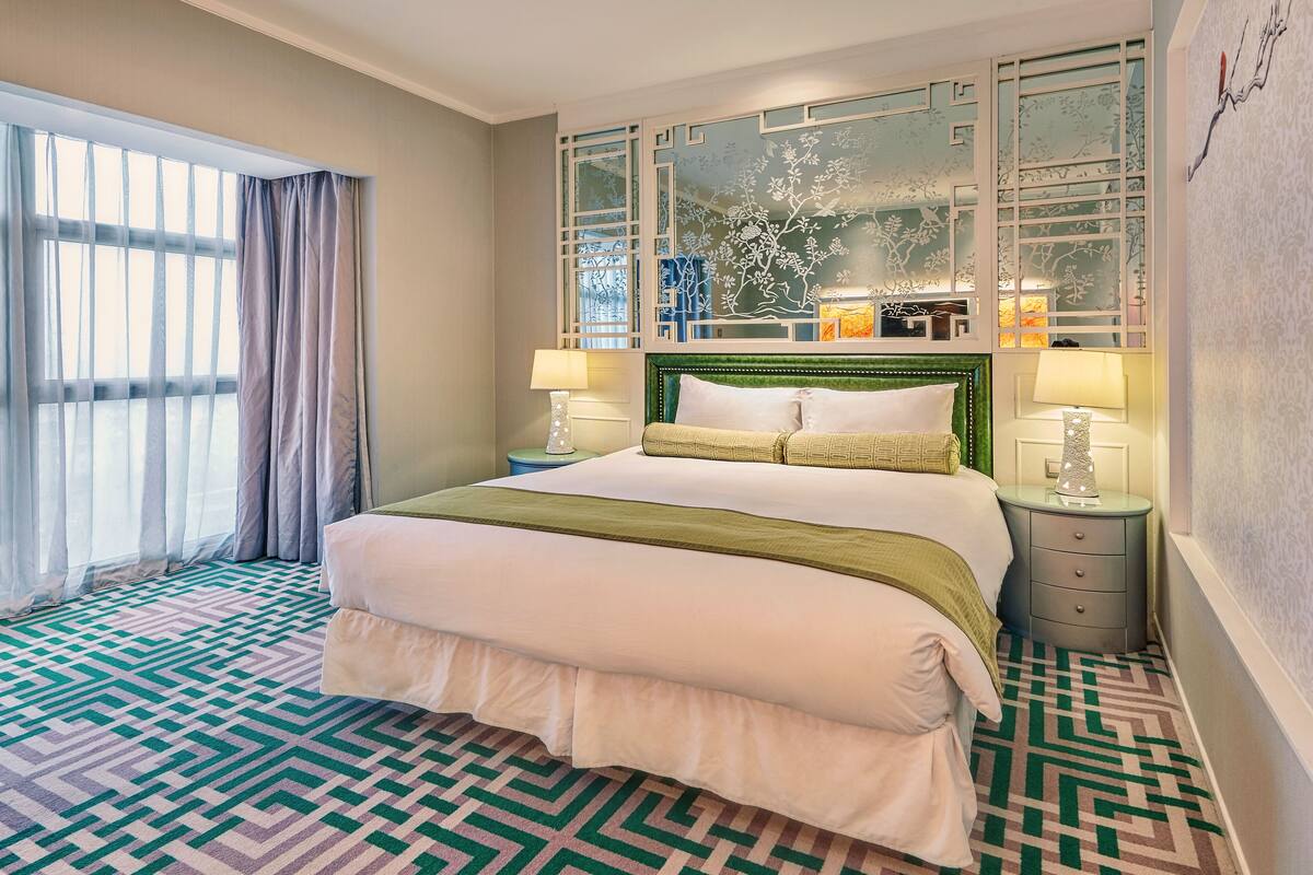 Una habitación de hotel con una cama grande, dos mesitas de noche con lámparas, un panel decorativo en la pared y una alfombra estampada.