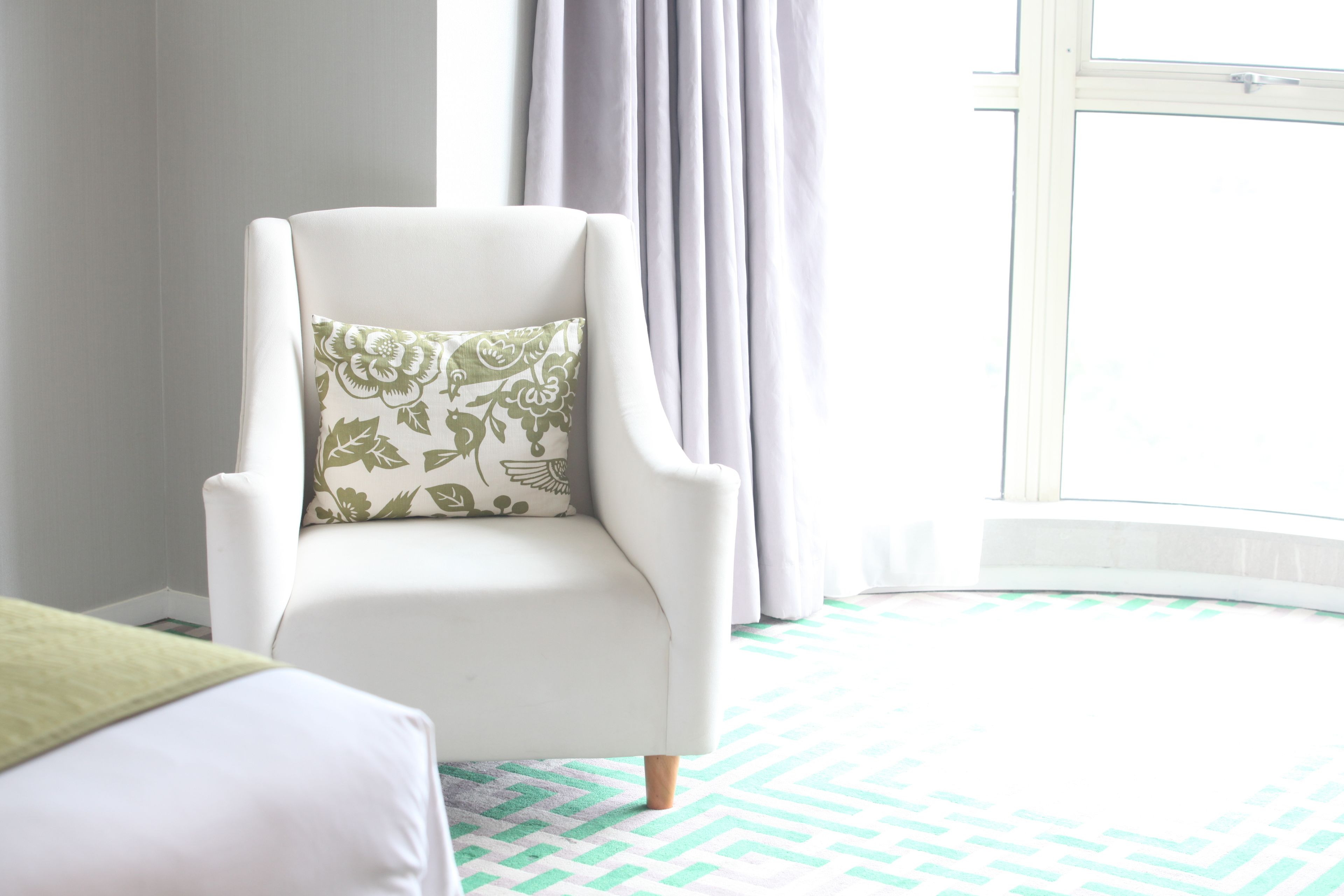 A white armchair with a floral pillow, positioned near a window with curtains, in a room with a patterned floor.