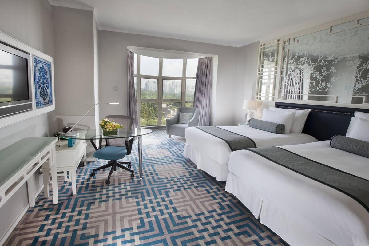 Habitación de hotel con dos camas, un escritorio con computadora, una silla y un ventanal con vista a la ciudad.