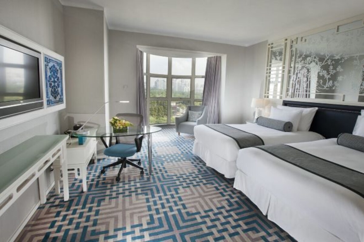 A hotel room with two beds, a desk, a chair, a TV, and a large window with a view of the city.