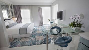 A modern hotel room with a bed, a glass desk, a chair, a TV, and a potted plant.