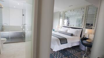 A modern hotel room with a glass shower enclosure, a large bed with a headboard, a bedside table with a lamp, and a patterned rug.