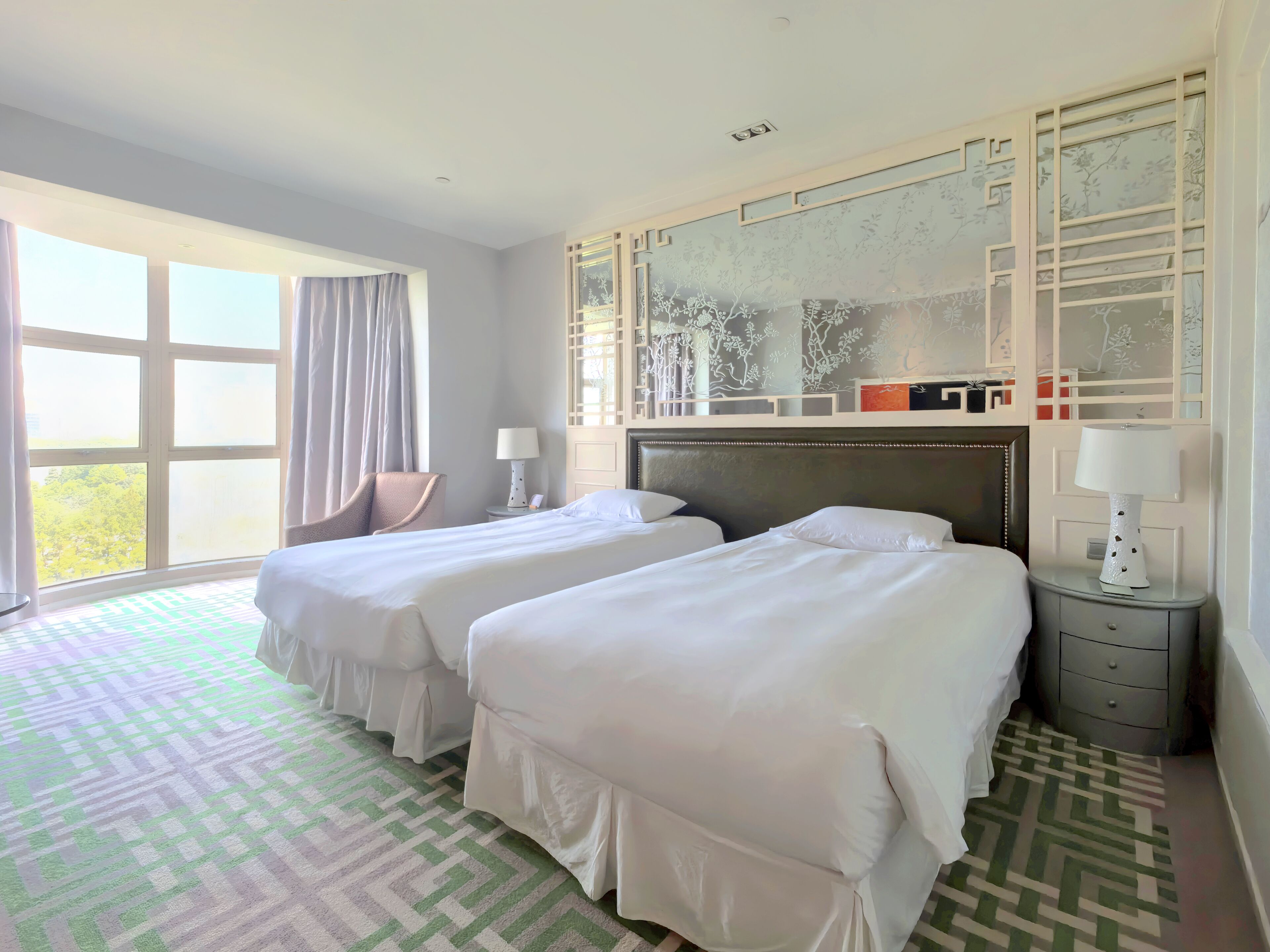 Une chambre d’hôtel avec deux lits, une grande fenêtre, une moquette à motifs et un panneau décoratif au mur.